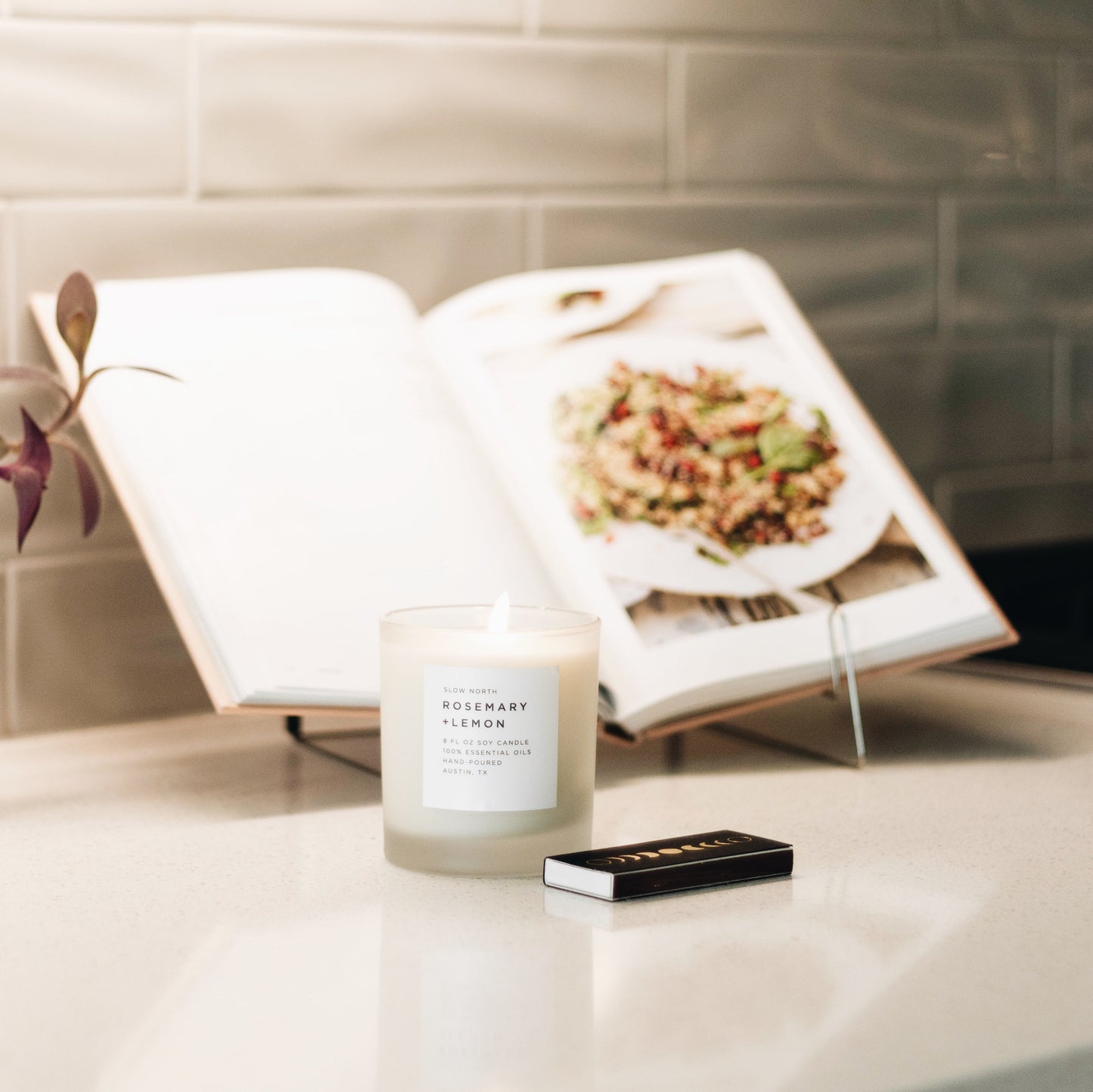 Lit soy wax candle in 8 ounce clear frosted tumbler with a black matchbook near base. Cookbook on stand in background on a counter. By Slow North