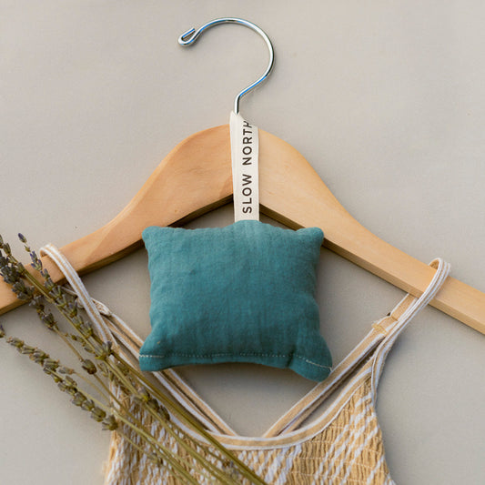 A Slow North cedar sachet hanging on a tan wooden hanger laying flat on a tan background with a yellow dress on the hanger with a lavender sprig.