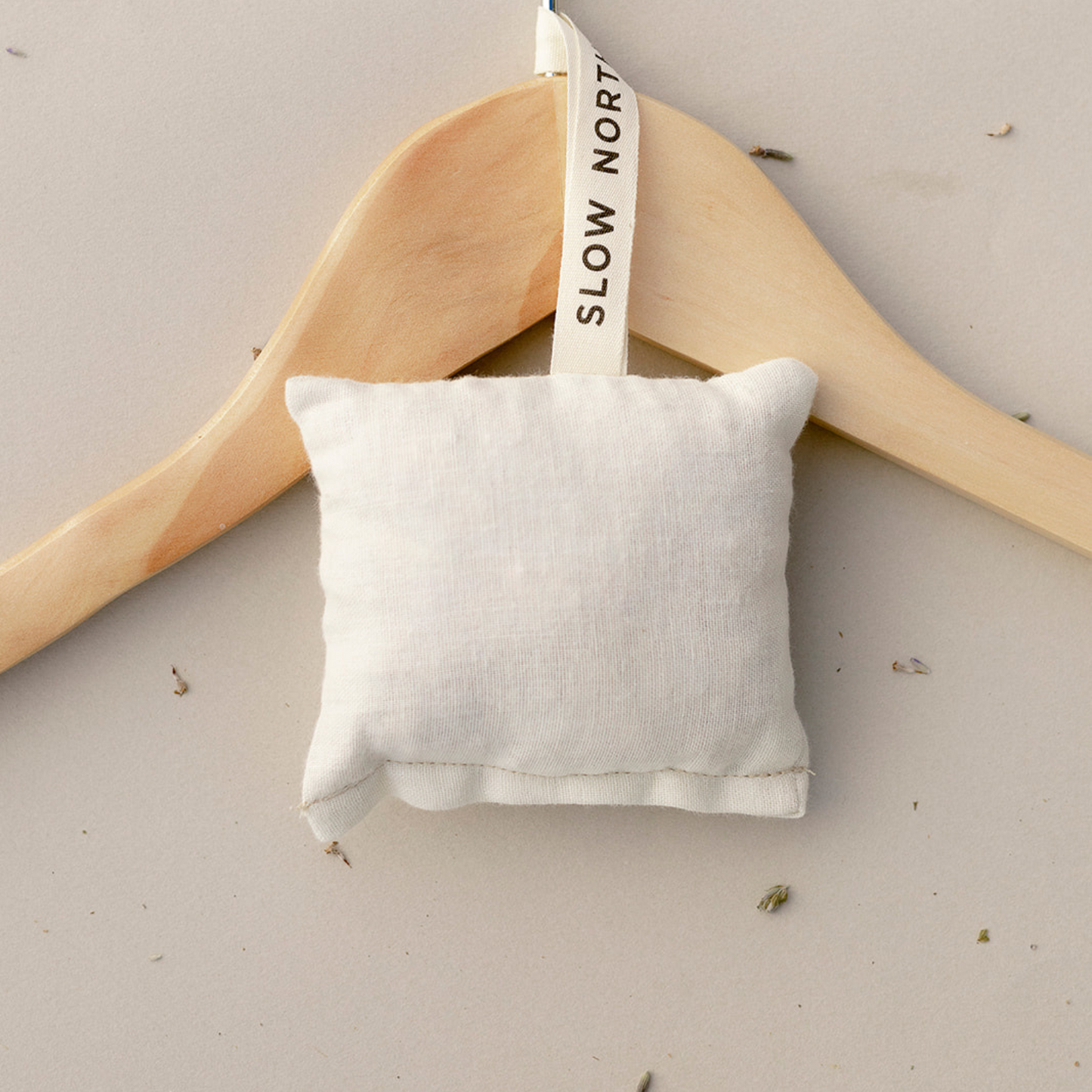 A Slow North lavender + cedar sachet hanging on a tan wooden hanger laying flat on a tan background with scattered loose lavender