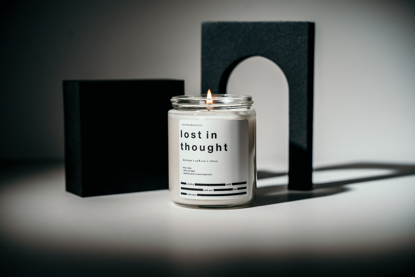 A postmodernform 'lost in thought' candle, which is white in a clear glass jar with a white label and black text containing the candle name, scent, and a themed poem, appears in front of artistic black shapes.