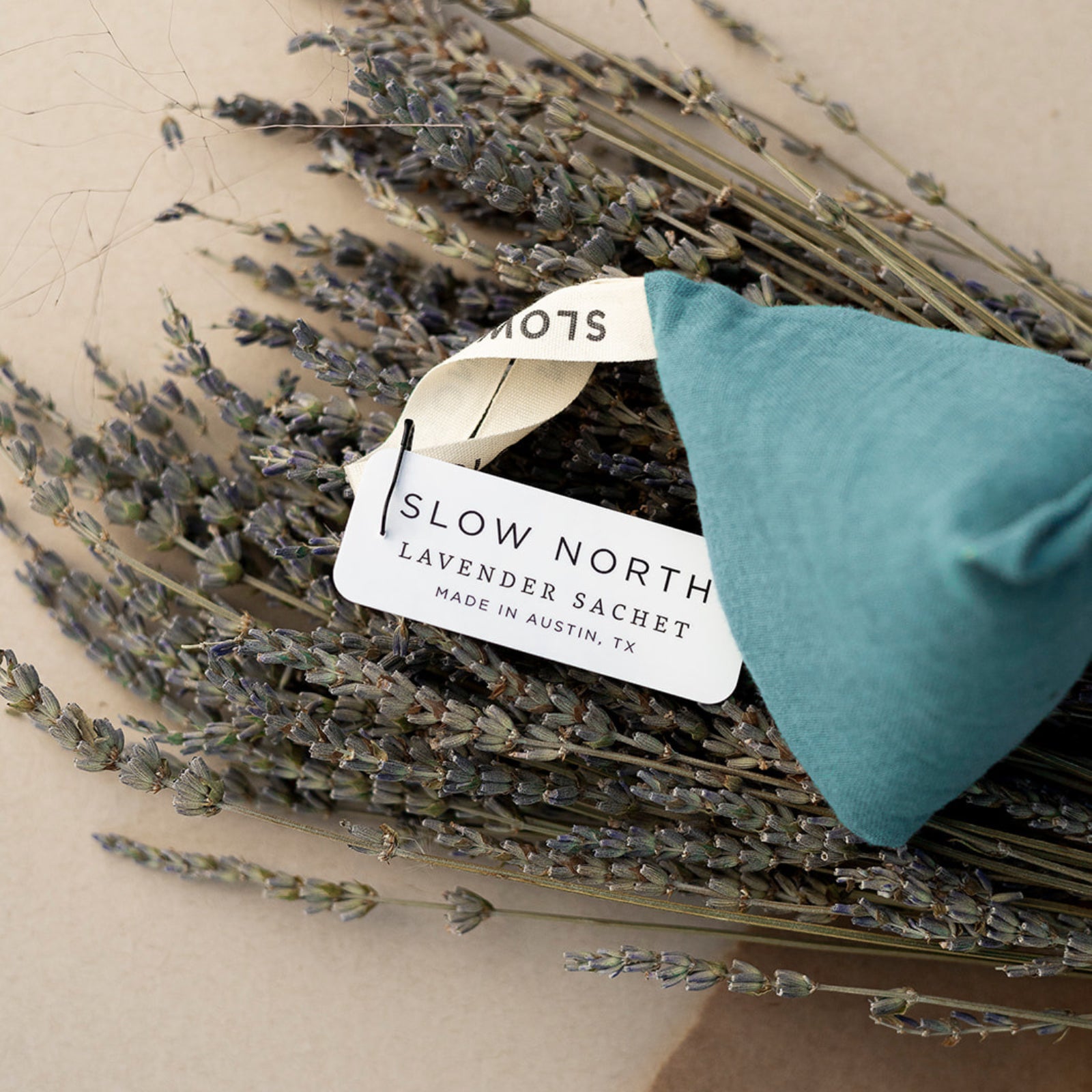 Lavender Sachet - Lagoon, a teal sachet prism laying flat on a bundle of lavender. The tag is visible. TEXT: SLOW NORTH LAVENDER SACHET Made in Austin, TX